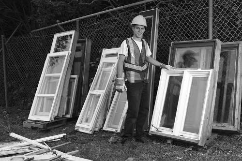 A male worker wearing a safety helmet and high-visibility vest stands outdoors on a patch of grass beside a six-foot wire fencing, next to a collection of wooden framed windows with glass panes leaning against the fence. The windows vary in size and design, with some featuring multiple panes separated by muntins, and have a light finish on their wooden frames. The frames exhibit a mix of textures, with smooth painted surfaces and natural wood grain visible on some. A few disassembled wooden window components are scattered on the ground in front. The background includes trees and foliage, suggesting a rural or suburban setting, potentially in or near Walthamstow, East London. The scene is lit with natural daylight, indicating typical daytime conditions suitable for rubbish removal and clearance work, as managed by Walthamstow House Clearance in the local area. This image depicts a professional rubbish removal process involving the collection and preparation of unwanted window frames for disposal or recycling, emphasizing the practical aspects of waste management services focused on property clearance.