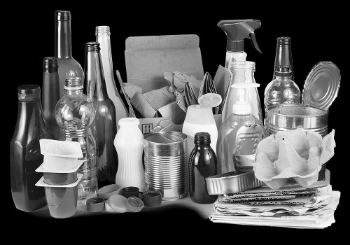 A collection of various discarded household items and waste materials arranged on a flat surface, including clear glass bottles, plastic containers, and packaging, alongside crumpled newspaper and paper towels, with some items stacked or leaning against each other. The bottles are in different shapes and sizes, primarily transparent with some opaque plastic, and some have caps or lids. The paper waste is piled neatly on one side, while other objects appear to be loosely grouped. The background is dark, emphasizing the contrast and textures of the waste items, which may be part of a rubbish clearance process carried out by Walthamstow House Clearance services, serving the E17 area nearby, with the scene set in an indoor environment or a collection point for rubbish removal.