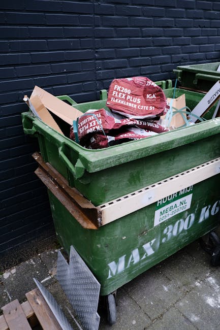 A street scene featuring a white industrial rubbish collection vehicle parked adjacent to a row of older multi-storey buildings with weathered facades, some displaying patches of peeling paint and exp